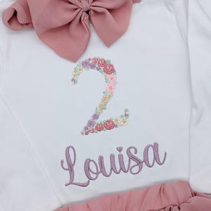 Könnte beinhalten: Weißes Langarm-Shirt mit einer rosa Schleife und einer floralen Stickerei der Zahl 2. Das Shirt hat auch den Namen "Louisa" in lila gestickt.