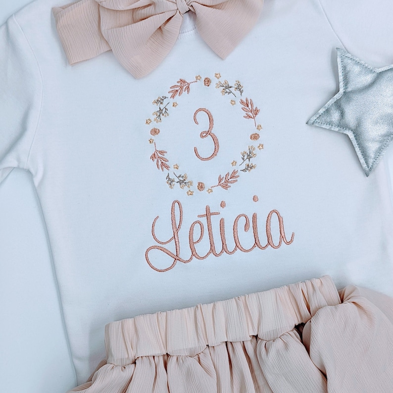 May include: A white onesie with a floral wreath embroidered around the number "3" and the name "Leticia" underneath. A pink bow and a silver star are also included in the image.