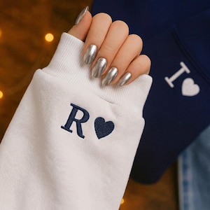 Puede incluir: Primer plano de una manga de sudadera blanca con la letra "R" y un corazón bordados en azul marino. El puño es blanco. En el fondo, una sudadera azul marino con el texto "I" y un corazón blanco.