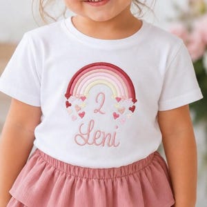 Könnte beinhalten: Ein weißes Kurzarm-T-Shirt mit einer Stickerei eines mehrfarbigen Regenbogens in Rot-, Rosa- und Gelbtönen. Unter dem Regenbogen ist die Zahl 2 in Rosa gestickt, gefolgt von dem Namen Leni in einer rosafarbenen Schreibschrift.