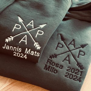 Op de afbeelding: Twee donkergroene hoodies met witte en zwarte geborduurde pijlen en de letters "PAP" op de voorkant. De bovenste hoodie heeft de tekst "Jannis Mats 2024" geborduurd onder de pijlen. De onderste hoodie heeft de tekst "Rosa Milo 2021 2024" geborduurd onder de pijlen.