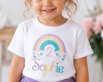 May include: A white short-sleeved t-shirt featuring an embroidered design of a pastel rainbow with small hearts and the number 2 in the center. Below the rainbow, the name Sophie is embroidered in a colorful script font on the front of the shirt.