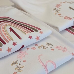 May include: Two white t-shirts with embroidered designs. The top shirt features a rainbow with a number one in the center and stars. The bottom shirt features a floral wreath with a number one in the center.