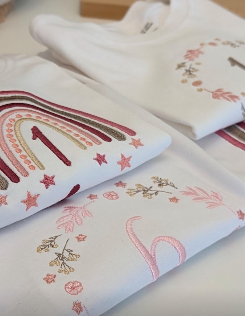 May include: Two white t-shirts with embroidered designs. The top shirt features a rainbow with a number one in the center and stars. The bottom shirt features a floral wreath with a number one in the center.