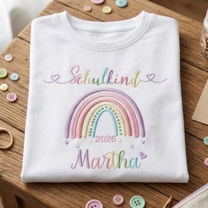 Könnte beinhalten: Ein weißes T-Shirt mit gesticktem Text, der Schulkind und Martha in einer bunten Schreibschrift zeigt. Zwischen den Namen befindet sich ein pastellfarbenes Regenbogenmotiv mit der Jahreszahl 2026 in der Mitte, verziert mit kleinen Herzen.