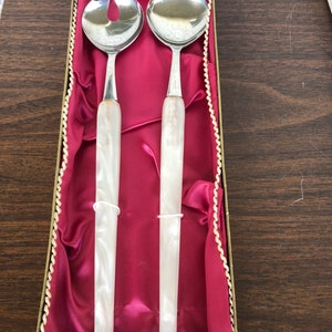 Könnte beinhalten: Ein Set aus zwei silbernen Salatbesteck mit weißen Perlmuttgriffen in einer roten Samtbox mit goldenen Verzierungen.