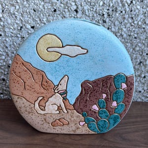 May include: A ceramic money box shaped like a desert scene with a coyote howling at the moon, a cactus, and mountains. The bank is painted in shades of blue, brown, green, and yellow.