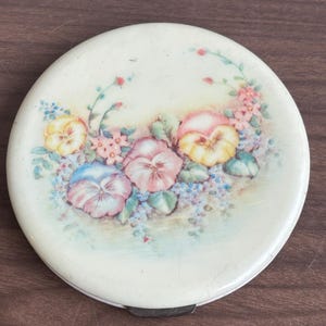 Op de afbeelding: Een ronde, crèmekleurige compact met een bloemmotief. De deksel toont een aquarel-achtige schildering van viooltjes in roze, geel en blauw, omringd door groene bladeren en kleine roze bloemen. Het heeft een metalen sluiting.