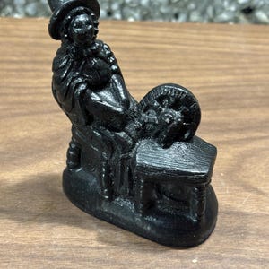 Può includere: Piccola statuina nera raffigurante una persona seduta a un filatoio. La figura indossa un cappello ed è seduta su una panca. Il filatoio è di fronte alla figura. La statuina è realizzata in un materiale scuro e solido.