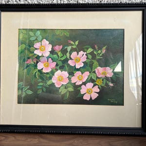 May include: A framed floral painting featuring pink flowers with yellow centers and green leaves. The artwork is set against a dark background and framed in a black, ornate frame. The painting is signed in the bottom right corner.
