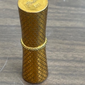May include: A gold-coloured lipstick case with a textured, scale-like pattern. The top of the case has the word "Renlor" and the number "581" engraved. The case has a cylindrical shape with a slightly flared base.