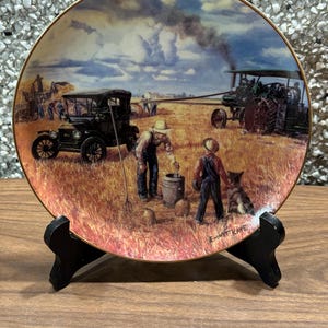 Puede incluir: Plato decorativo que representa una escena rural con un coche antiguo, un tractor y figuras en un campo. El plato tiene un borde dorado y se exhibe en un soporte negro. La obra de arte incluye un perro y el texto "Emmett Kaye".