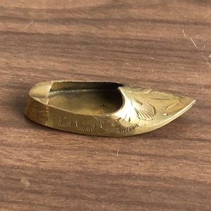 May include: A small, gold-colored, decorative shoe-shaped ashtray. The ashtray is intricately engraved with a floral pattern.