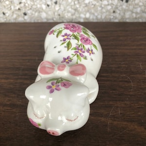May include: A white ceramic pig figurine with pink accents and purple and pink floral designs. The pig is lying down with its eyes closed and has a pink bow around its neck.