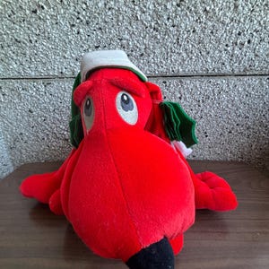May include: A plush red dragon toy with a white hat and green holly leaves. The dragon has large, round eyes, a black nose, and a soft, cuddly appearance. This festive toy is perfect for holiday decor or as a gift.
