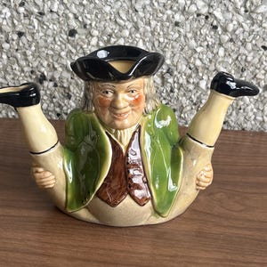 Puede incluir: Una tetera de cerámica con forma de hombre con las piernas en el aire. La tetera presenta un sombrero negro, una chaqueta verde y un chaleco marrón. La cara del hombre sonríe con mejillas rosadas. Un artículo decorativo único.