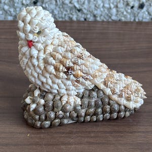 Peut inclure: Une figurine décorative d'oiseau faite de divers coquillages. L'oiseau présente une tête et un corps blancs, avec des coquillages bruns et beiges sur les ailes et le dos. La base est composée de petits coquillages gris, et une petite coquille rouge forme le bec.