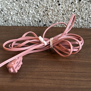 May include: A coiled, pale pink electrical cord with a matching plug. The cord is secured with a white twist tie and a rubber band. The plug has two flat prongs and a rectangular shape.
