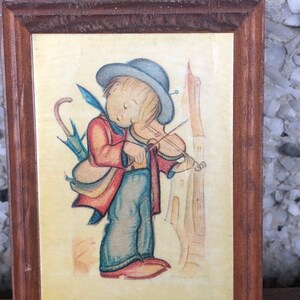 May include: A framed illustration of a young person playing a violin. The person is wearing a red jacket, blue trousers, and a blue hat. The illustration is set against a yellow background with a tall building in the background.