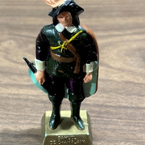 May include: A miniature figurine of Samuel de Champlain, a historical figure, stands on a rectangular base. The figure wears a dark green coat, black hat with a feather, and a cape. The base is inscribed with the name "SAMUEL DE CHAMPLAIN".