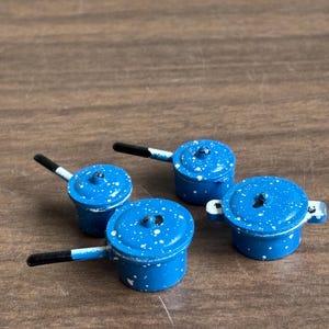May include: Set of four blue enamel miniature pots with white speckles. The pots have black handles and are arranged on a wooden surface.