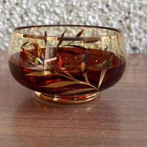 Puede incluir: Un pequeño cuenco redondo de vidrio rojo con un borde dorado y un diseño de hojas doradas. El cuenco está sobre una superficie de madera.