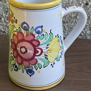 Puede incluir: Taza de cerámica blanca con borde y asa amarillos. La taza está decorada con un diseño floral en tonos rojos, amarillos, verdes y azules.