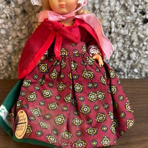 May include: A vintage doll wearing a red dress with a yellow and white floral pattern, a red cape, and a white lace bonnet. The doll has blonde hair and blue eyes. The doll is labelled "Romby Danish Design."