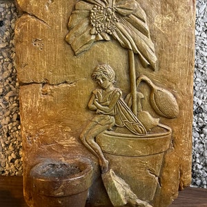 Op de afbeelding: Een houten wandplaquette met een gegraveerde fee die in een pot zit met een bloem die eruit groeit. De plaquette heeft een rustiek bruin en groen kleurenschema.