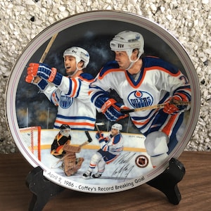 May include: A commemorative porcelain plate featuring a hockey scene with three players in action. The plate depicts a player scoring a goal, another player skating with the puck, and a goalie in the net. The plate is decorated with a blue and orange color scheme and includes the text "April 2, 1986 - Coffey's Record Breaking Goal".