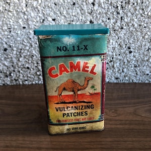 Pode incluir: Uma caixa de lata vintage com uma tampa turquesa e um rótulo colorido com a silhueta de um camelo. O rótulo diz "CAMEL VULCANIZING PATCHES FOR TUBELESS TIRES AND TUBES 40 OBLONG" e "NO. 11-X".
