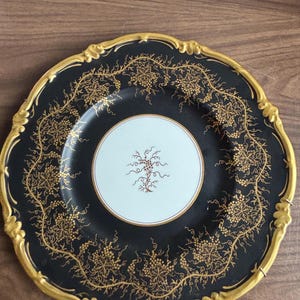 May include: A round decorative plate featuring a black center and rim with an intricate gold-colored vine and grape pattern. The plate has a scalloped edge with a raised gold-colored border and a white circular center with a small brown floral motif.