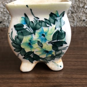 Puede incluir: Un macetero de cerámica blanca con un diseño floral con flores azules, verdes y amarillas. El macetero tiene tres patas y una abertura cuadrada en la parte superior.