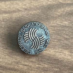 May include: A round, silver-toned pin with the text "CELEBRATE SASKATCHEWAN" and the years "1905-1980" encircling a stylized emblem. The pin is set against a wooden surface.