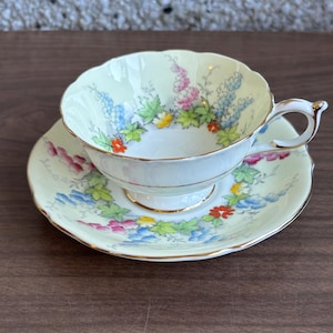 May include: A vintage teacup and saucer set. The cup and saucer are pale yellow with a floral design in blue, pink, and red. The edges are scalloped and trimmed with gold. A classic tea set.