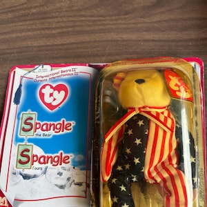 May include: A Ty Beanie Baby bear named Spangle, in its original packaging. The bear has a yellow head and a body decorated with red and white stripes and navy blue fabric with white stars. The packaging includes the text "Spangle the Bear".