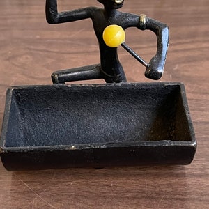Puede incluir: Una figura de metal negro de una persona tocando una batería, con baquetas y parches amarillos. La figura está arrodillada sobre una bandeja rectangular. La figura tiene detalles dorados en los brazos.