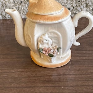 Könnte beinhalten: Eine kleine, weiße Teekanne aus Keramik mit braunem Deckel und floralem Design. An der Seite der Teekanne befindet sich eine kleine weiße Figur eines Mädchens, das Blumen hält.