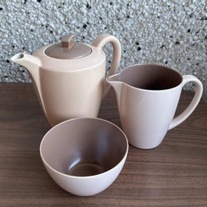 Può includere: Un set da tè in tre pezzi in una palette di colori neutri. Il set comprende una teiera con coperchio, una lattiera e una piccola ciotola. L'interno della ciotola e della lattiera è marrone scuro, mentre l'esterno è beige chiaro.