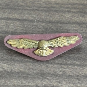 Op de afbeelding: Een vintage-stijl speld met een goudkleurige adelaar met uitgespreide vleugels. De adelaar is gecentreerd op een bordeauxrode, schildvormige basis. De speld heeft een gedetailleerd ontwerp en een klassieke uitstraling.