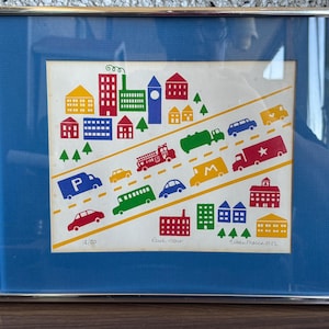 May include: A framed print depicting a vibrant cityscape with buildings, trees, and vehicles. The artwork includes a fire engine, a bus, and various cars. The print is set against a blue background and framed in silver.