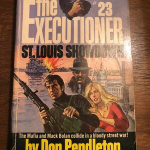 May include: A paperback book cover featuring a graphic of three people holding guns. The book title is "The Executioner" and the subtitle is "St. Louis Showdown". The author is Don Pendleton. The book is number 23 in a series.