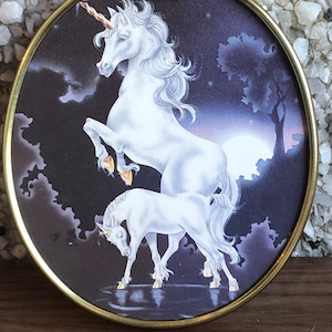 May include: A round, gold-framed wall decor featuring a white unicorn and its foal standing in a moonlit forest. The unicorn has a gold horn and a flowing mane and tail.