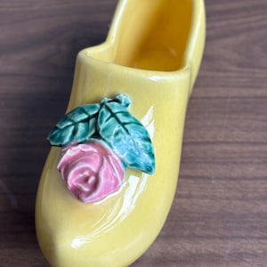 May include: A yellow ceramic clog-shaped vase with a pink rose and green leaves. The vase is a bright yellow colour and has a smooth, glossy finish. The rose and leaves are three-dimensional and add a decorative touch.
