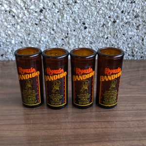 May include: Four brown glass shot glasses with the words "Kahlua Bandido" printed on them. Each glass has a different image of a cartoon bandit with a sombrero and a bottle of Kahlua. The text on the glasses reads "A blend of Kahlua liqueur and imported premium gold tequila."