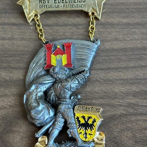 May include: A silver metal pin with a depiction of a knight holding a flag with a red and yellow castle. The pin has a gold chain and a gold banner with the text "1978 5.INT. VOLKSWANDERTAG RSV EDELWEISS OFFENBURG-FESSENBACH".