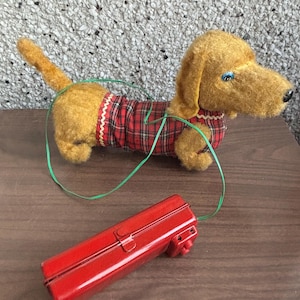 Pode incluir: Um brinquedo vintage de dachshund de corda com uma camisa xadrez vermelha e preta. O cão de brinquedo é castanho e tem um cordão verde ligado a uma chave de corda de metal vermelha.