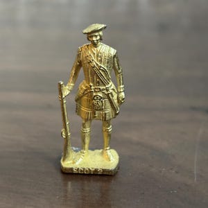 May include: A small, gold-colored figurine of a Scottish soldier in traditional attire, including a kilt, beret, and rifle. The figurine is standing on a rectangular base with the text "SCOT 2". The figure is approximately 2 inches tall.