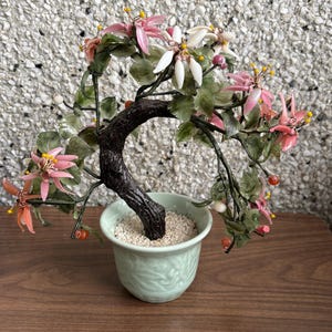 May include: A miniature bonsai tree with a dark brown trunk and delicate pink and white flowers. The tree is planted in a light green ceramic pot filled with small white stones. The bonsai is on a wooden surface.