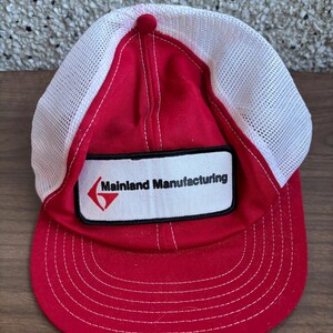 Pode incluir: Boné de caminhoneiro vermelho e branco com um remendo preto que diz "Mainland Manufacturing" em letras brancas com um logotipo de seta vermelha.
